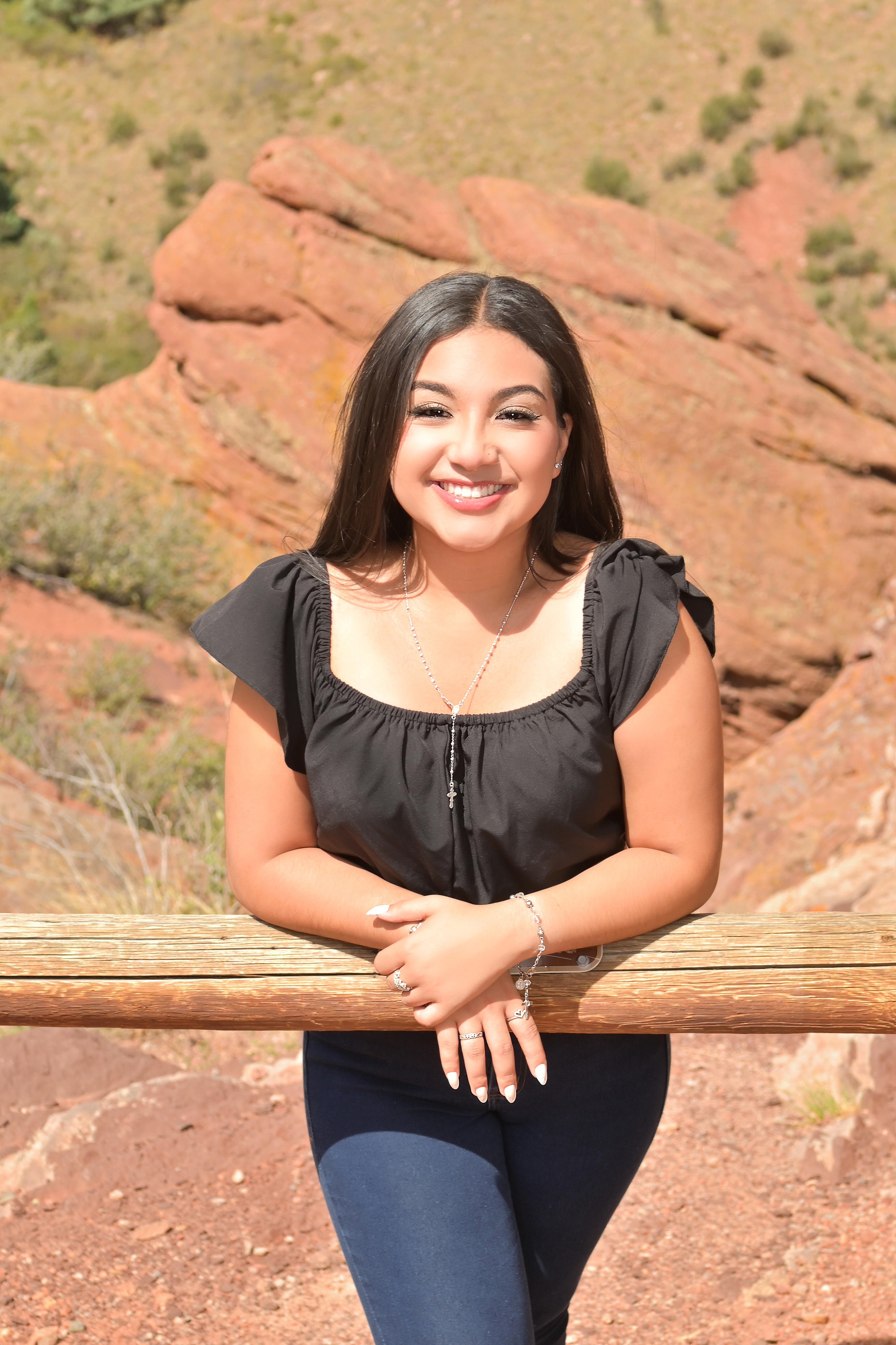 A female student is outside in the mountains leaning up against a wooden post smiling towards the camera.